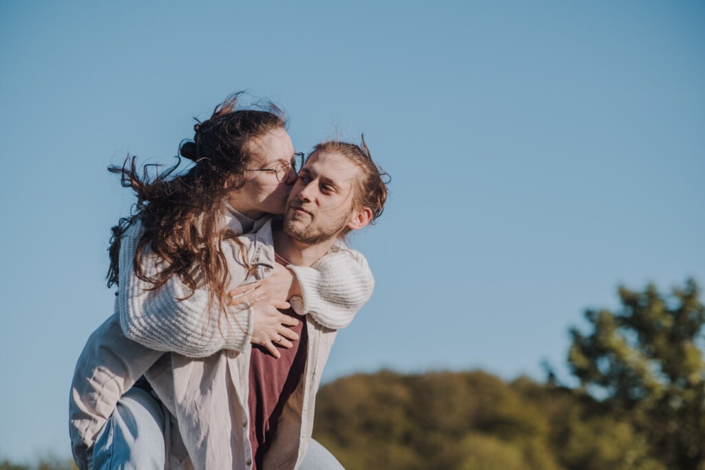 Paar-Fotoshooting Travemünde Jawortmomente Ein Mann trägt eine Frau huckepack, während sie ihm einen Kuss auf die Wange gibt; beide tragen Freizeitkleidung vor einem klaren blauen Himmel.